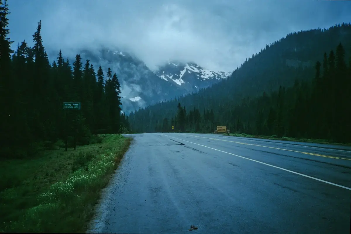 Nasse Passstraße durch Nadelwald, Wolken hängen an schneebedeckten Gipfeln