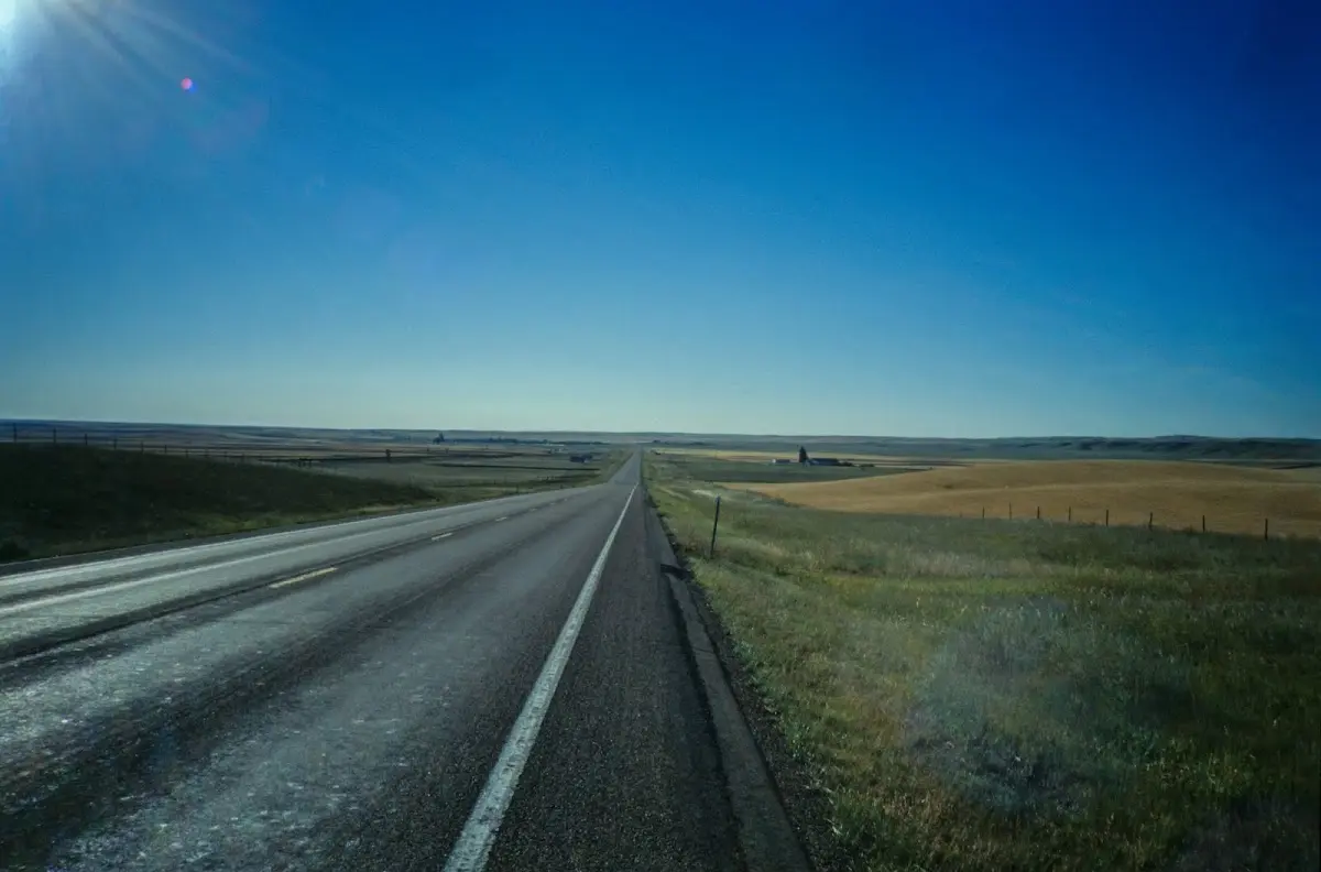 Schnurgerade Straße durch die Prärie bis zum Horizont, rechts ein Weizenfeld, Gegenlicht