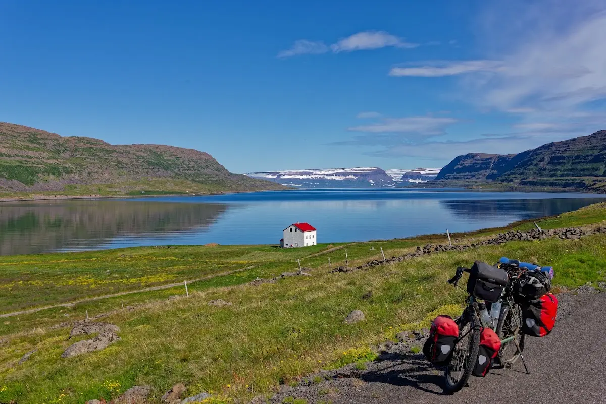 Reiserad mit roten Packtaschen am Straßenrand über einem ruhigen Fjord in den Westfjorden, ein weißes Haus mit rotem Dach am Ufer